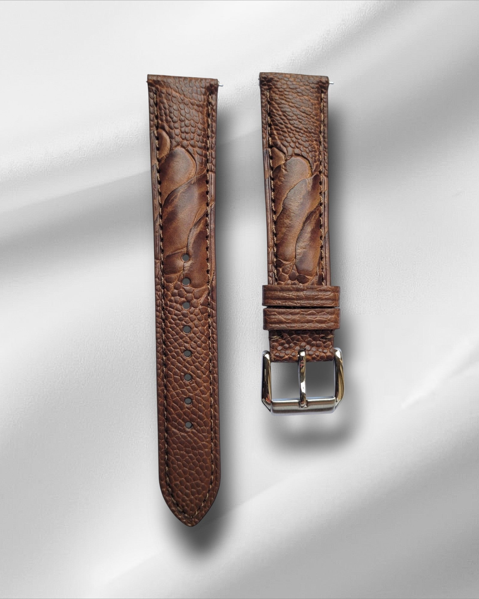 A Tan ostrich leg leather watch strap with a steel tang buckle, displayed against a white background.